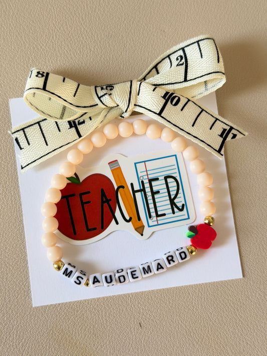 A handmade teacher-themed beaded bracelet displayed on a white card. The bracelet features pale peach-colored beads, a red apple charm, a small red apple with green leaves, and letter tiles spelling 'MS.